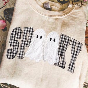 May include: A beige crewneck sweatshirt featuring the word SPOOKY appliqued across the front. The letters S, P, K, and Y are made of black and white gingham fabric, while the two O's are replaced by white lace ghost shapes with black eyes.