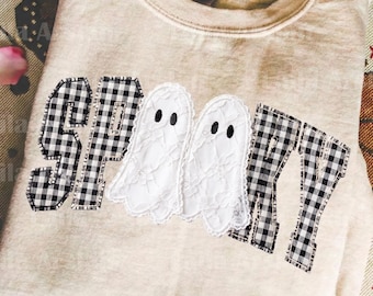 May include: A beige crewneck sweatshirt featuring the word SPOOKY appliqued across the front. The letters S, P, K, and Y are made of black and white gingham fabric, while the two O's are replaced by white lace ghost shapes with black eyes.
