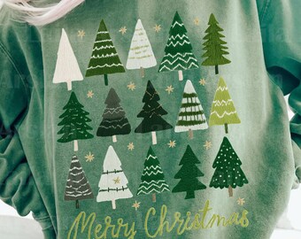May include: Green sweatshirt with a festive design featuring multiple Christmas trees in various shades of green and white, with gold star accents. The words "Merry Christmas" are written in a cursive font across the bottom.