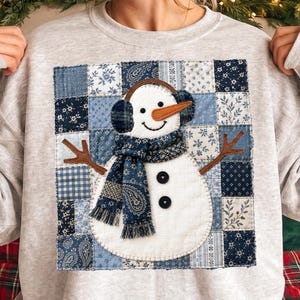 Könnte beinhalten: Ein hellgraues Sweatshirt mit Rundhalsausschnitt und einem großen quadratischen Aufnäher im Patchwork-Design aus verschiedenen blau-weißen Blumen- und Geometriemustern. Ein weißer Schneemann mit braunen Stockarmen, blauem Paisleyschal und Ohrenschützern.