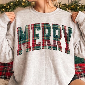 Può includere: Una felpa girocollo grigio chiaro con la parola MERRY sul petto in grandi lettere in stile blocco. Le lettere sono riempite con un motivo in tessuto scozzese rosso, verde e giallo e presentano una finitura sfilacciata lungo i bordi.