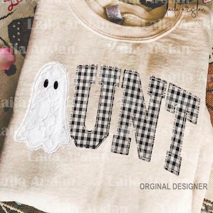 May include: A beige crewneck sweatshirt featuring a white lace ghost appliqué with black eyes and the letters U, N, and T in a black and white buffalo plaid pattern. The text is stitched onto the front of the garment in a collegiate font.