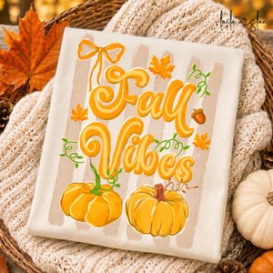 以下が含まれることがあります： オレンジ色のスクリプトで「Fall Vibes」と書かれたクリーム色のTシャツ。カボチャ、葉、ドングリで飾られています。シャツにはベージュの縦縞があり、居心地の良いクリーム色のセーターの上に表示されています。