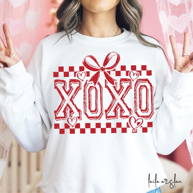 Puede incluir: Sudadera blanca con un patr&oacute;n de cuadros rojos y un lazo rojo. La palabra "XOXO" est&aacute; escrita en rojo con un aspecto desgastado.