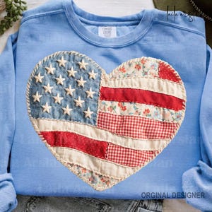 May include: Light blue sweatshirt featuring a heart-shaped American flag design. The flag is made of denim, red and white patterned fabric, and white stars. The sweatshirt has the words "Comfort Colors" on the tag.
