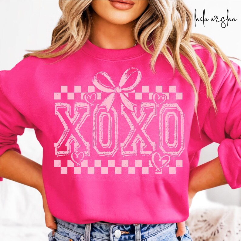 Puede incluir: Una sudadera rosa con un patr&oacute;n de cuadros blancos y la palabra "XOXO" en una fuente rosa, delineada. Un lazo rosa con un coraz&oacute;n a cada lado est&aacute; por encima de la palabra.