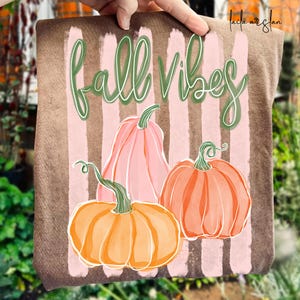 May include: A brown rectangular sign with vertical pink stripes and the words "fall vibes" in green script. Three watercolor pumpkins in shades of orange and pink are featured below the text, evoking an autumn theme.
