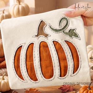 Puede incluir: Un artículo de tela color crema con una gran aplicación central de calabaza. La calabaza está hecha de segmentos de tela naranja quemado con un tallo marrón y una enredadera verde con una hoja, todo delineado con costuras visibles.
