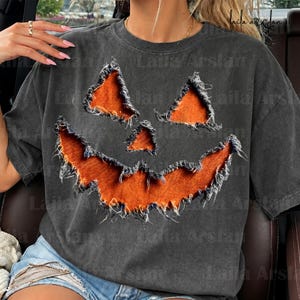 May include: A charcoal gray short-sleeve t-shirt featuring a large, distressed jack-o'-lantern face graphic. The pumpkin eyes, nose, and jagged mouth appear as orange fabric cutouts with frayed, raw edges layered onto the dark gray shirt fabric.