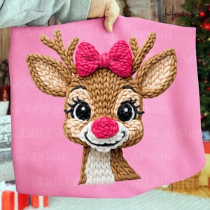 Könnte beinhalten: Ein rosa Stoff mit einem gestrickten Rentiergesicht. Das Rentier hat braunes Fell, große schwarze Augen mit langen Wimpern, eine rote Nase und eine rosa Schleife. Das Rentier lächelt. Der Hintergrund ist ein zartes Rosa.