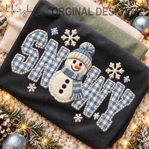 May include: A dark gray sweatshirt featuring the embroidered word SNOWY in blue and white gingham fabric. A central snowman applique wears a blue knit hat and scarf. White embroidered snowflakes surround the text and snowman design.