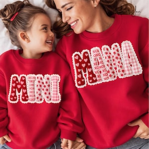 May include: Red sweatshirts with the words "MAMA" and "MINI" in a decorative font. The "MAMA" sweatshirt has a heart and floral design, while the "MINI" sweatshirt has a heart and gingham design. Both sweatshirts have a white lace trim.