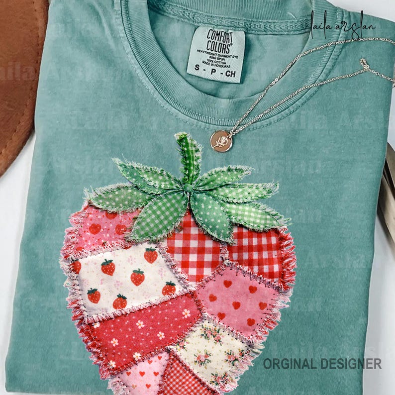 May include: A light teal Comfort Colors t-shirt with a patchwork strawberry design. The strawberry is made of various red and pink fabrics, including gingham and floral patterns. A silver necklace with a small pendant rests on the shirt.