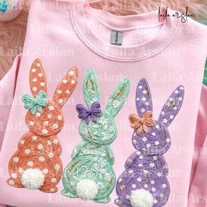 Könnte beinhalten: Rosa Sweatshirt mit drei gestickten Osterhasen in Orange, Mintgrün und Lila. Jeder Hase hat eine Schleife und weiße Punkte. Das Sweatshirt ist von der Marke Gildan.