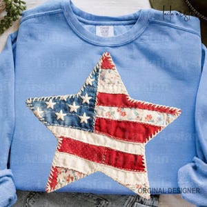 May include: Light blue Comfort Colors sweatshirt featuring a large, star-shaped patch. The patch is designed to resemble the American flag, with blue denim and white star accents, and red and white striped and floral fabric. The sweatshirt has a crew neck.