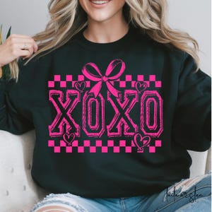 Puede incluir: Sudadera negra con un patr&oacute;n de cuadros rosas, un lazo rosa y la palabra "XOXO" en letras rosas.