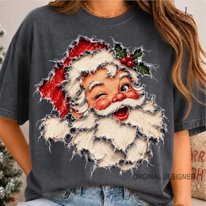 May include: A charcoal gray short-sleeve t-shirt featuring a distressed, layered-look graphic of a winking Santa Claus wearing a red hat with holly leaves and berries. The design has a frayed, textured appearance across the chest area.