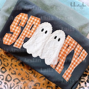 Könnte beinhalten: Ein dunkelgraues T-Shirt mit dem Wort SPOOKY, appliziert aus orange-weißem Vichy-Stoff. Die beiden Buchstaben O sind durch weiße Spitzengeister mit schwarzen gestickten Augen ersetzt. Das T-Shirt liegt auf einem Stoff mit Leopardenmuster.