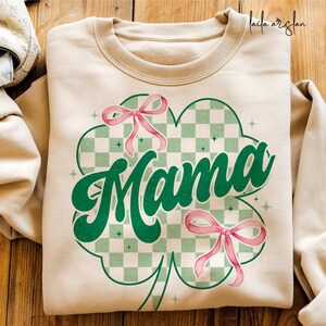 May include: A beige sweatshirt with a green four-leaf clover design. The clover has a checkered pattern and the word "Mama" in a green retro font. There are two pink bows on the clover.