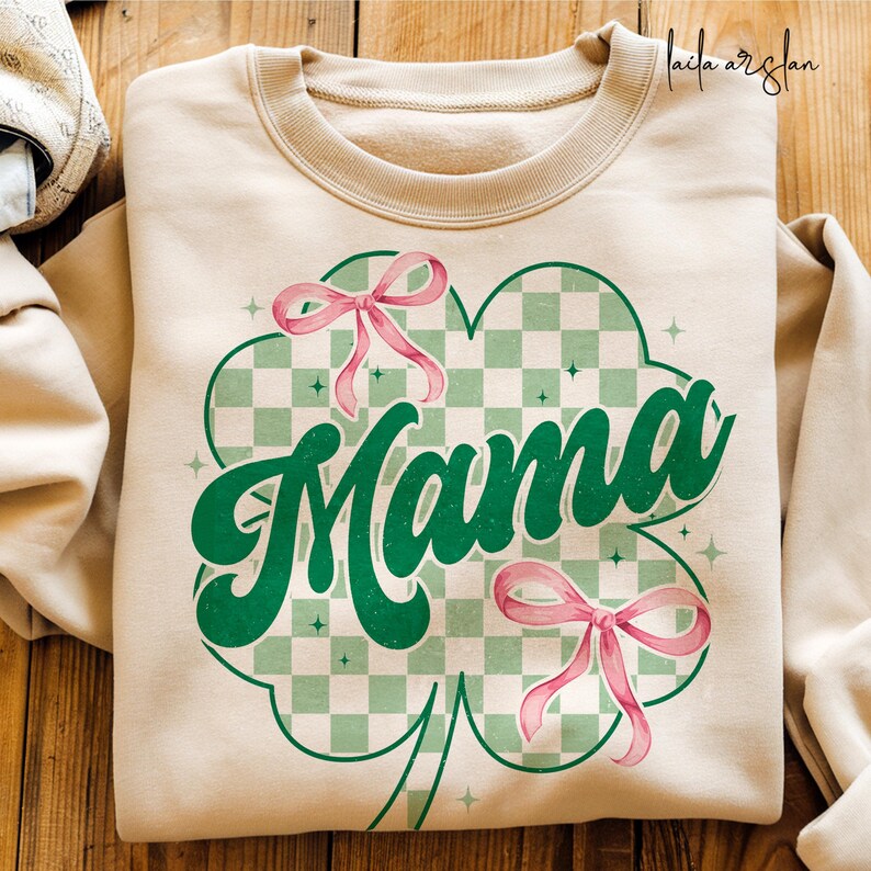 May include: A beige sweatshirt with a green four-leaf clover design. The clover has a checkered pattern and the word "Mama" in a green retro font. There are two pink bows on the clover.