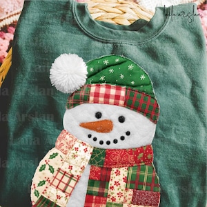Könnte beinhalten: Ein grünes Sweatshirt mit einem Schneemann-Applikation. Der Schneemann hat eine weiße Bommelmütze, eine grün-rot karierte Mütze, eine Karottennase und einen Patchwork-Schal in Rot, Grün und Creme. Das Gesicht des Schneemanns hat schwarze Punktaugen und ein gesticktes Lächeln.