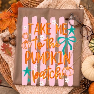 Peut inclure: Un t-shirt marron avec le texte "Take Me To The Pumpkin Patch" en orange et blanc. Le motif comprend des rayures roses, des citrouilles, des étoiles et des fleurs. Un collier doré avec un pendentif citrouille et des lunettes sont également visibles.