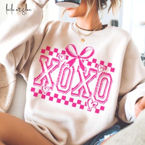 Puede incluir: Una sudadera de color crema con un gr&aacute;fico XOXO rosa. El gr&aacute;fico presenta un patr&oacute;n de cuadros, un lazo rosa y corazones.