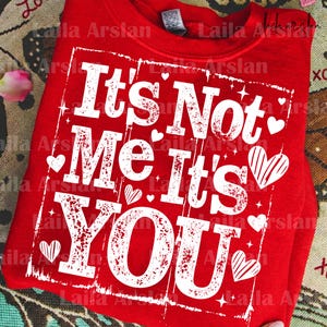 Op de afbeelding: Rode sweatshirt met de tekst "It's Not Me It's YOU" in witte letters. Het ontwerp bevat kleine hartjes en sterren. Een Valentijnsdag thema top.
