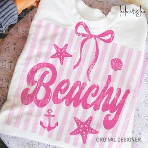 May include: White sweatshirt with a pink striped design and the word "Beachy" in a distressed font. The design includes a bow, starfish, a seashell, and an anchor. The sweatshirt is a casual, beach-themed apparel item.