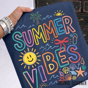 May include: A navy blue pillow with the words "SUMMER VIBES" in colorful, outlined letters. The design includes a smiling sun, palm tree, waves, seashells, and a flip-flop. The words "ORIGINAL DESIGNER" are at the bottom.