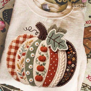 May include: A beige crewneck sweatshirt featuring a large, textured pumpkin design created with punch needle embroidery. The pumpkin is divided into vertical sections with varied patterns including orange gingham, floral motifs, white daisies, and cable knit.