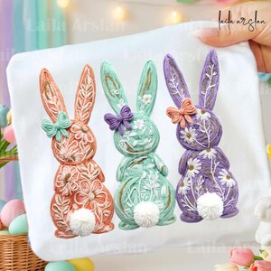 Puede incluir: Una tela blanca con tres conejos decorativos de Pascua. Los conejos están hechos de hilo y presentan detalles florales y lazos. Los conejos son en tonos naranja, verde menta y morado. Cada conejo tiene una cola de pompón blanca.