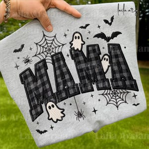 May include: A light gray sweatshirt featuring the word MAMA embroidered in a black and white buffalo plaid pattern. The design includes embroidered white ghosts, black spiderwebs, black spiders, and black bats scattered across the fabric.