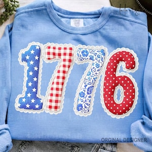 Może przedstawiać: Błękitna bluza z numerem "1776" naszytym z przodu. Numery są w czerwone, białe i niebieskie wzory, w tym gwiazdy, kratę i kwiatowe wzory. Bluza ma napis "Comfort Colors".