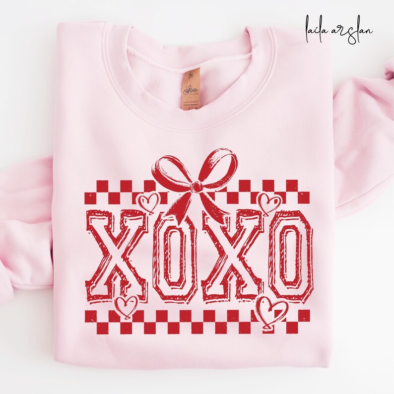 Puede incluir: Sudadera rosa con un gr&aacute;fico XOXO rojo. El gr&aacute;fico presenta un lazo rojo, un patr&oacute;n a cuadros y corazones.