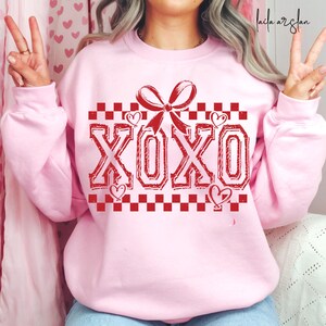 Puede incluir: Sudadera rosa con un patr&oacute;n de cuadros rojos y un lazo rojo con el texto "XOXO" en rojo.