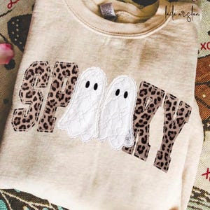 Könnte beinhalten: Ein beiges Sweatshirt mit Rundhalsausschnitt und dem Wort SPOOKY in Buchstaben aus Leopardenmuster-Stoff. Die beiden O's sind durch weiße Spitzen-Geister-Applikationen mit schwarzen gestickten Augen ersetzt. Das Sweatshirt hat einen Rippkragen.