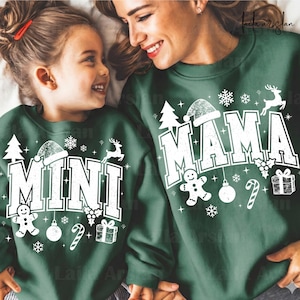 May include: Two people wearing matching green sweatshirts with the words "MINI" and "MAMA" in white, festive lettering. The sweatshirts feature Christmas-themed graphics like a Santa hat, snowflakes, and a gingerbread man.