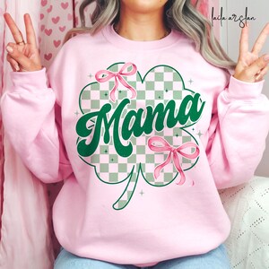 May include: Pink sweatshirt with a green shamrock design and the word "Mama" in a retro font. The shamrock has a checkered pattern and pink bows.