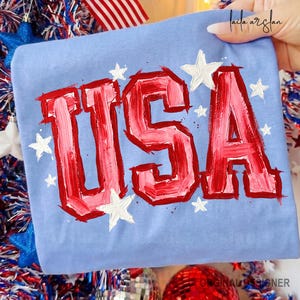 May include: A light blue t-shirt with the word "USA" in red and pink brushstrokes, surrounded by white stars. The shirt is folded, with the text prominently displayed. The design has a patriotic theme, suitable for Independence Day or other national holidays.