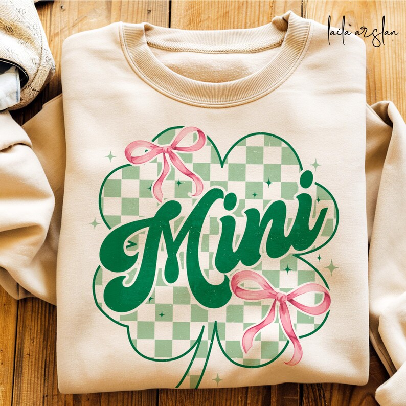 May include: A beige sweatshirt with a green shamrock design. The shamrock has a checkered pattern and the word "Mini" in green letters. There are pink bows on the shamrock.