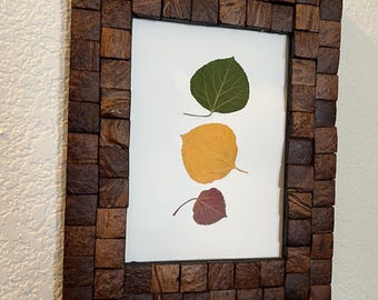May include: A rectangular wooden picture frame with a mosaic-like design. The frame surrounds a white background with three pressed leaves: green, yellow, and red. The frame is made of small, dark brown wooden blocks.