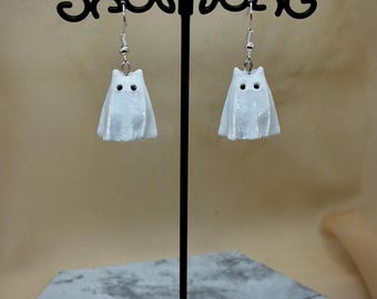 May include: White ghost-shaped earrings with black eyes and a draped design. The earrings hang from silver-colored hooks. The earrings are displayed on a black metal earring stand with a decorative top and base.