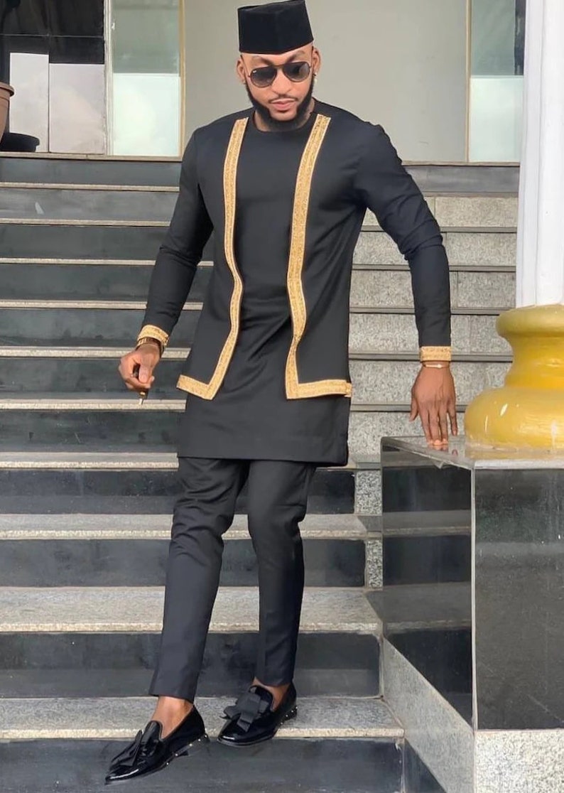 May include: A man wearing a black and gold traditional African outfit with a black cap and sunglasses. He is standing on a set of stairs.