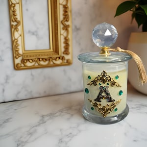 May include: A decorative glass candle jar with a faceted crystal lid and gold tassel. The jar is adorned with gold filigree, emerald-green gems, and a black letter "A". The candle inside has a pale yellow hue.
