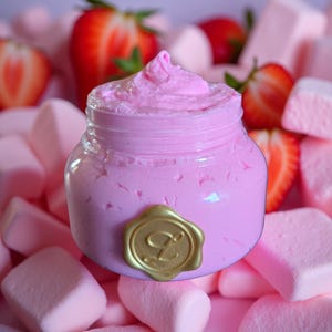 May include: A small glass jar filled with pink, whipped cream-like substance, topped with a swirl. The jar has a gold seal with a letter "L" on it. The jar is surrounded by pink marshmallows and fresh strawberries.