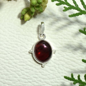 May include: A silver pendant with a red gemstone set in an oval shape. The pendant has four small silver accents on the sides.