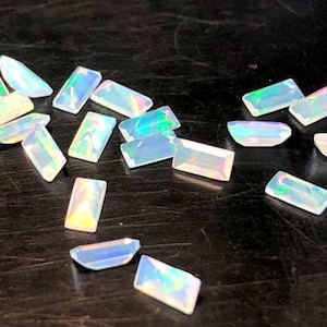 Puede incluir: Una colección de piedras preciosas de ópalo blanco iridiscente de forma rectangular. Las piedras preciosas están dispersas sobre una superficie de madera oscura.