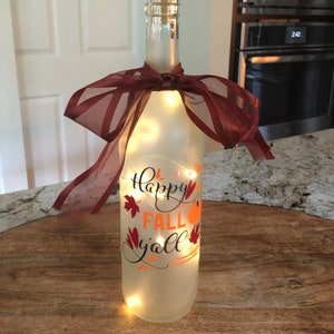 May include: A frosted glass wine bottle with a cork stopper and a burgundy ribbon. The bottle is decorated with the words "Happy Fall y'all" and fall-themed graphics. Small lights are inside the bottle, illuminating the text and design.