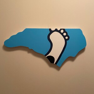 May include: A blue and white outline of the state of North Carolina with a white footprint inside. The footprint is outlined in black and has a black dot in the center.